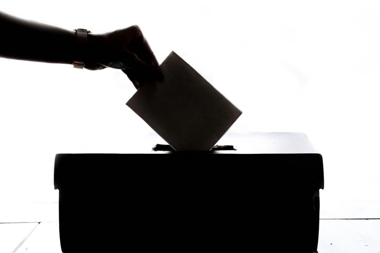 A hand drops a ballot into a ballot box in silhouette