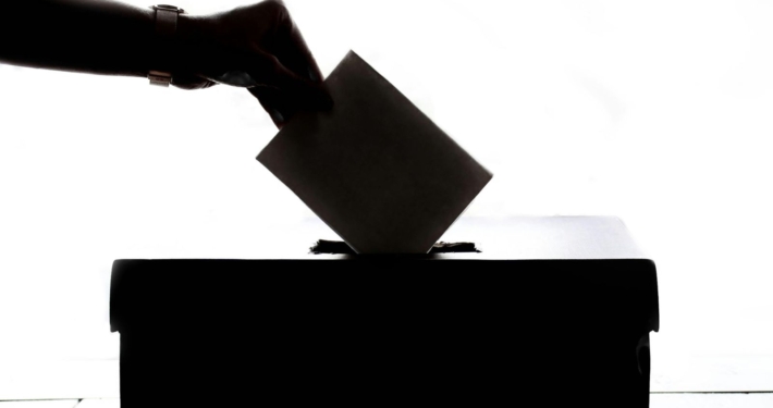A hand drops a ballot into a ballot box in silhouette